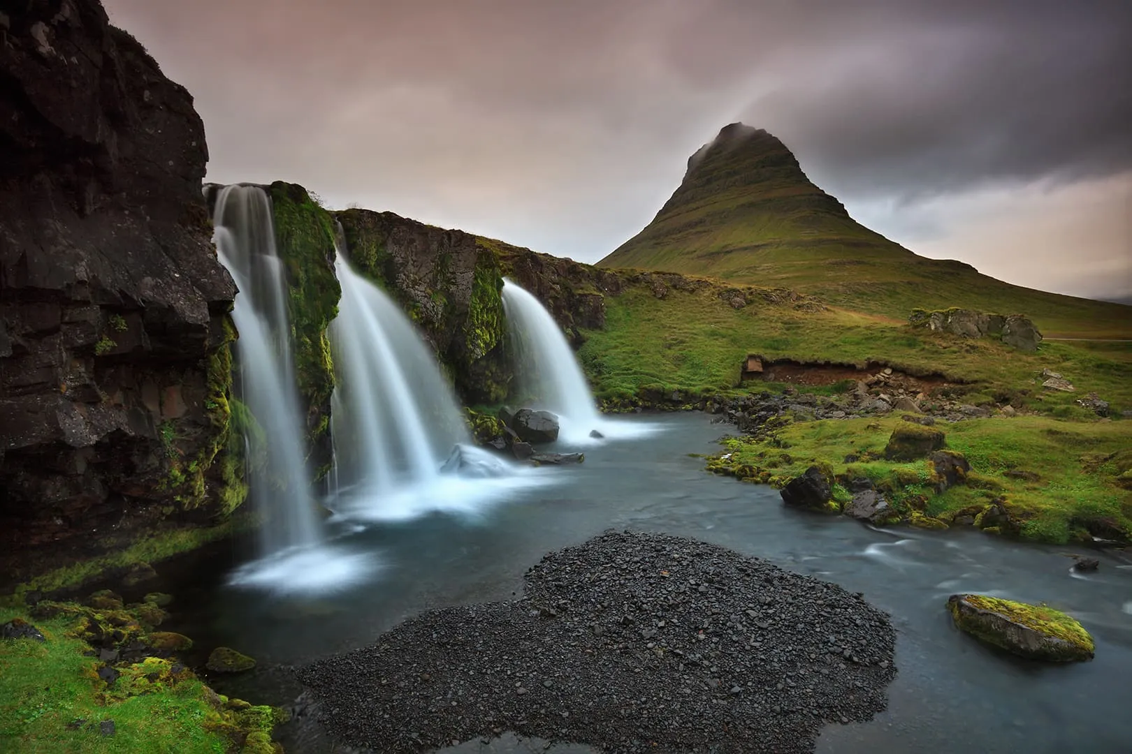 ภูเขา Kirkjufell ทรงกรวยกับน้ำตกสามสายไหลผ่านทุ่งหญ้าเขียว